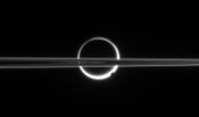 Saturn's rings cut across an eerie scene that is ruled by Titan's luminous crescent and globe-encircling haze, broken by the small moon Enceladus, whose cryovolcanos are dimly visible at its south pole. North is up. Imaged by Cassini in 2006.