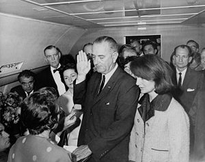 Lyndon B. Johnson being sworn in aboard Air Force One by Federal Judge Sarah T. Hughes, following the assassination of John F. Kennedy. To the left of Johnson is Jacqueline Kennedy, widow of Kennedy; to his right is Mrs. Lady Bird Johnson, and sitting down near the airplane window is Jack Valenti, founder of the MPAA.