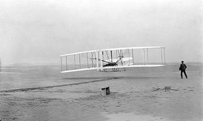 First flight of the Wright Flyer I, 17 December 1903, Orville piloting, Wilbur running at wingtip. Photo by John T. Daniels of the Kill Devil Hills Life Saving Station, using Orville's tripod-mounted camera