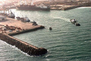 Aerial view of the Port of Mogadishu in 1992. Three cargo ships, large, medium and small sized vessels are moored to the docks. A tugboat is heading out of the port.