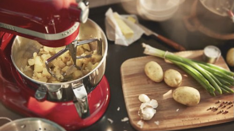 a red kitchenaid with potatoes in it sits on a kitchen counter next to a cutting board of scallions, potatoes, and garlic