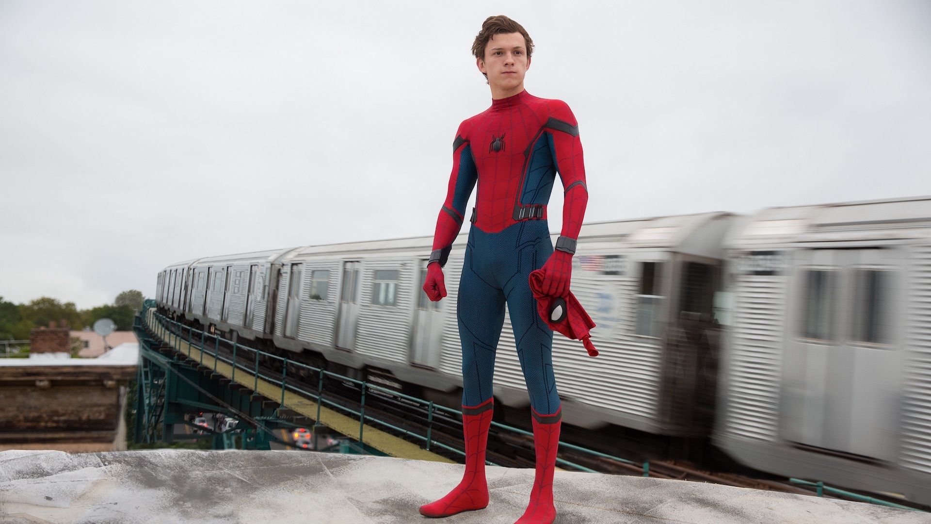 Tom Holland as Peter Parker, standing on a roof and wearing the Spider-Man suit, except for the mask which he holds in his hand.. A blurry New York City subway train speeds by in the background.