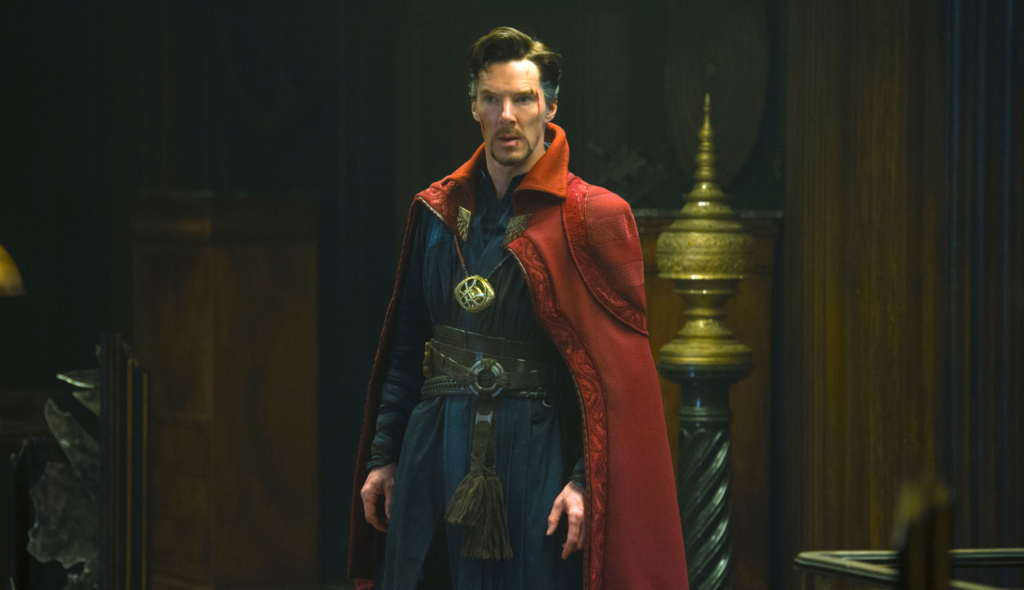 Dr. Stephen Strange (Benedict Cumberbatch) in a dark room, wearing his blue superhero outfit and signature red, collared cape. His face bears several cuts and scrapes.