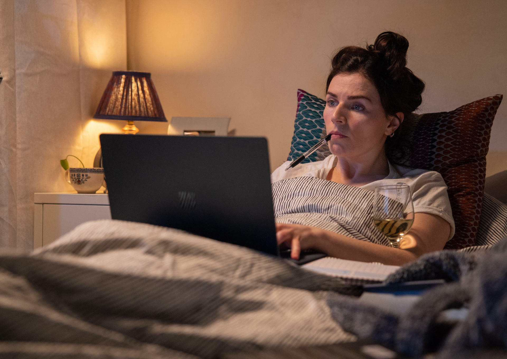 A picture of Aine (Aisling Bea) sitting in bad with her laptop and a glass of wine in "This Way Up."