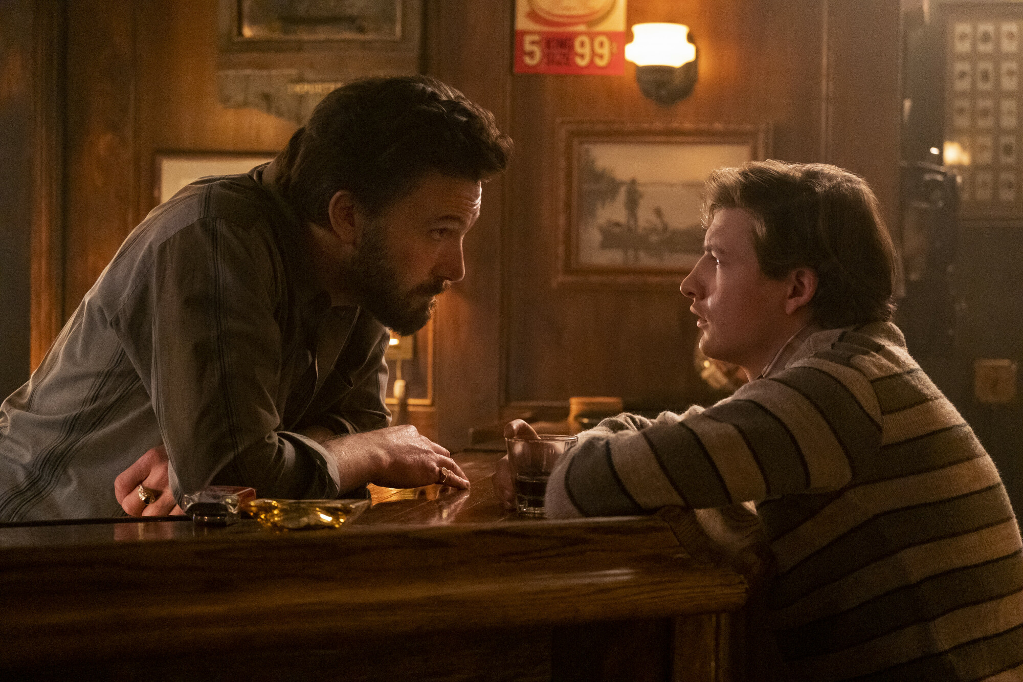 A bearded bartender leans over a bar toward a young man. 