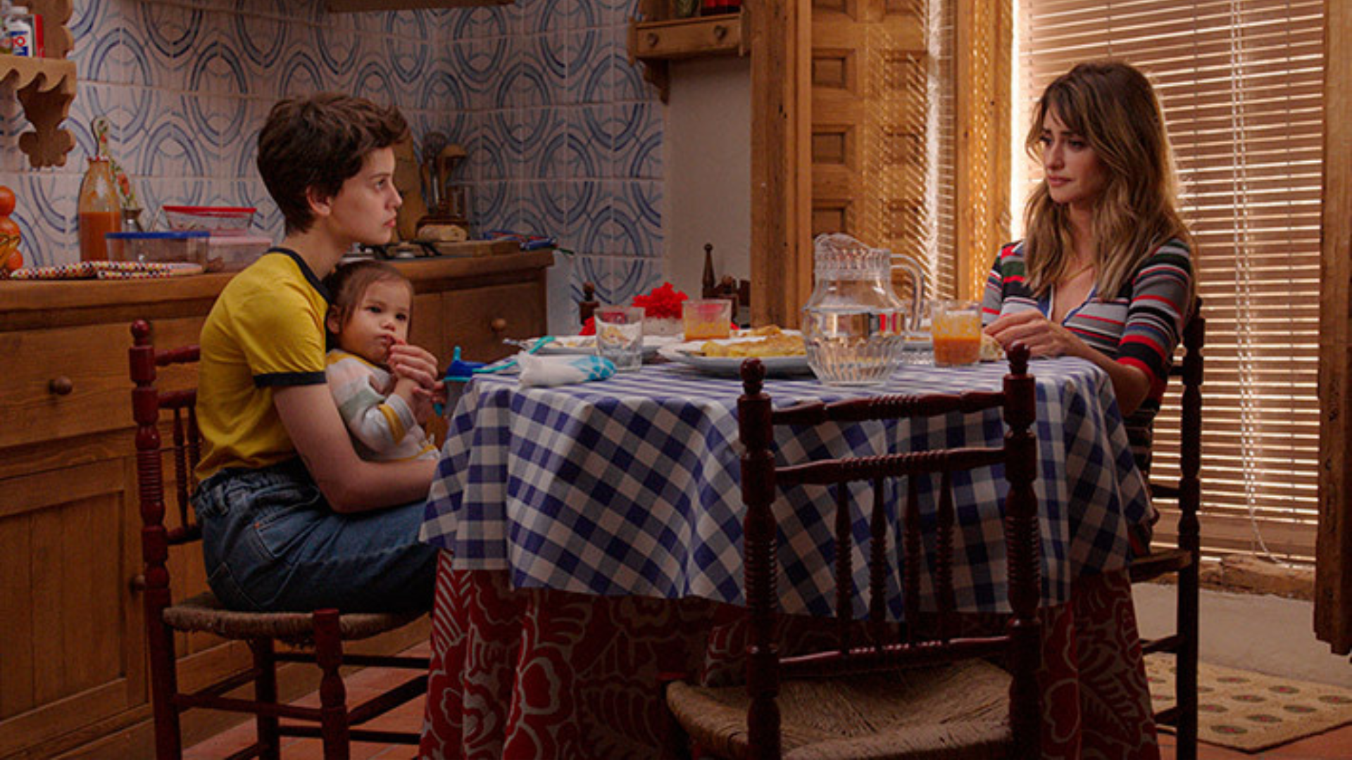 Two women sit at a kitchen table, the younger of the two has a baby on her lap.
