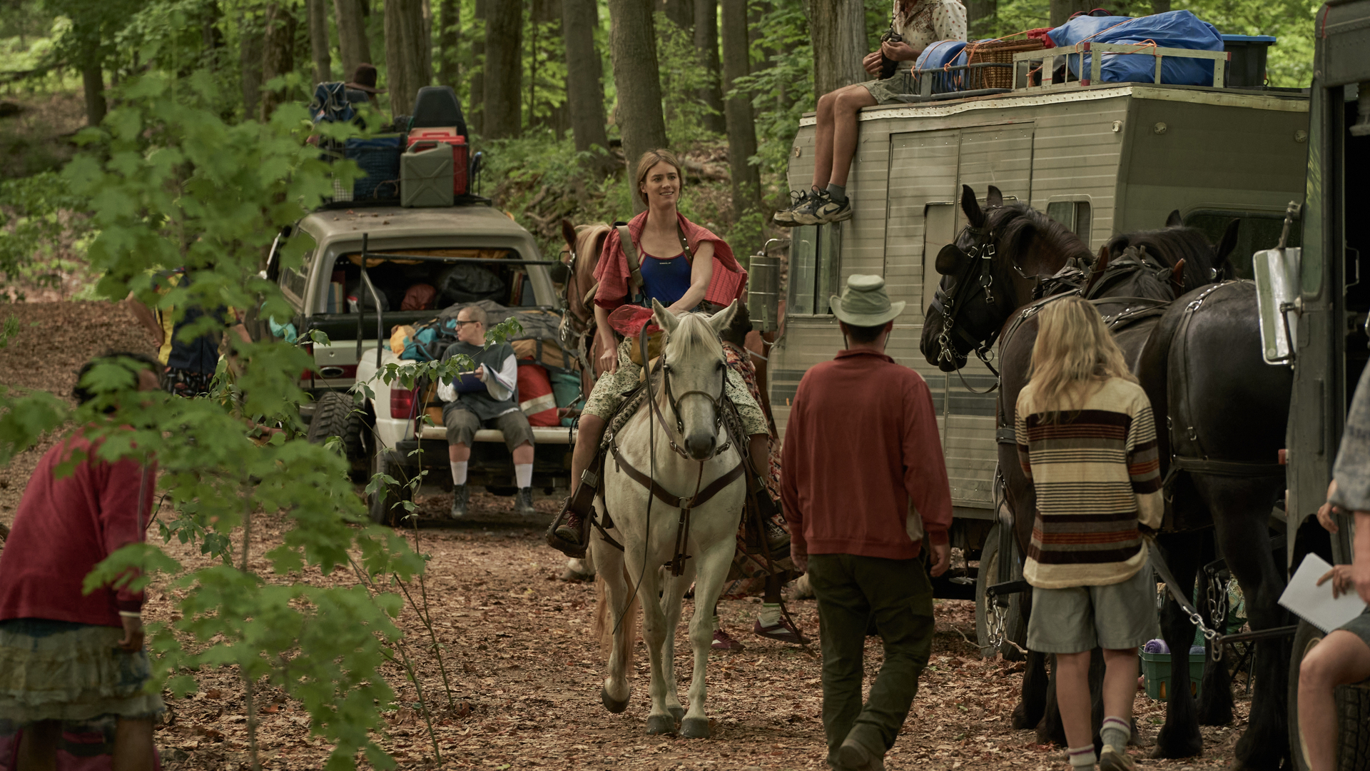 A woman rides a horse through a forest alongside other travelers in horse-drawn cars and trailers