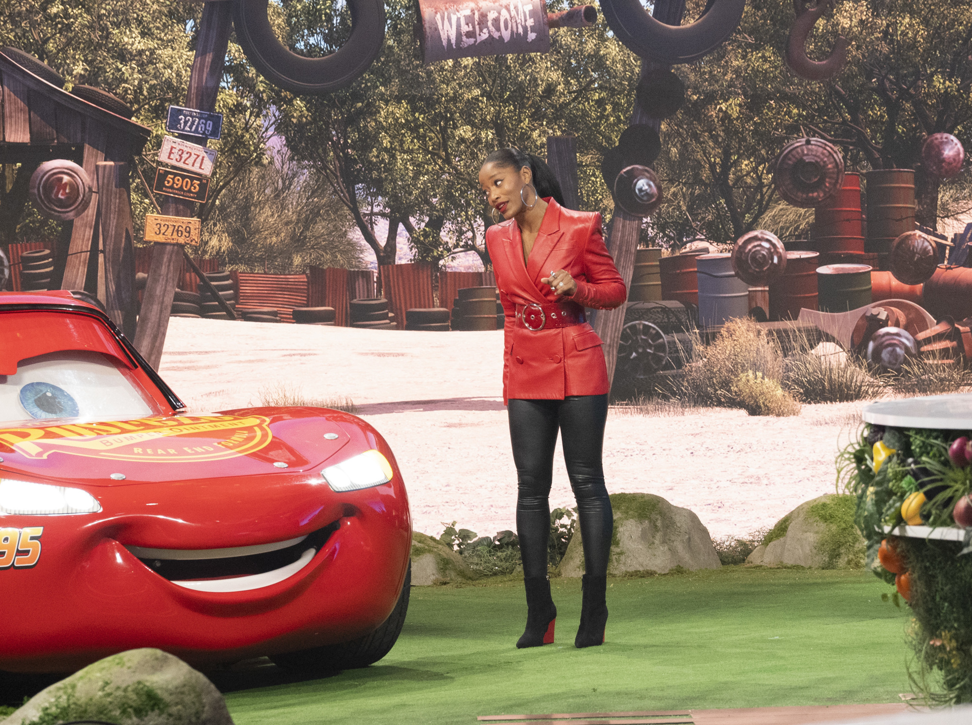 A still from the Disney+ reality competition series, "Foodtastic." Host Keke Palmer, wearing a belted red leather overcoat, black leggings, and black high-heeled boots, stands on a Cars-themed set next to a recreation of Cars star Lightning McQueen.