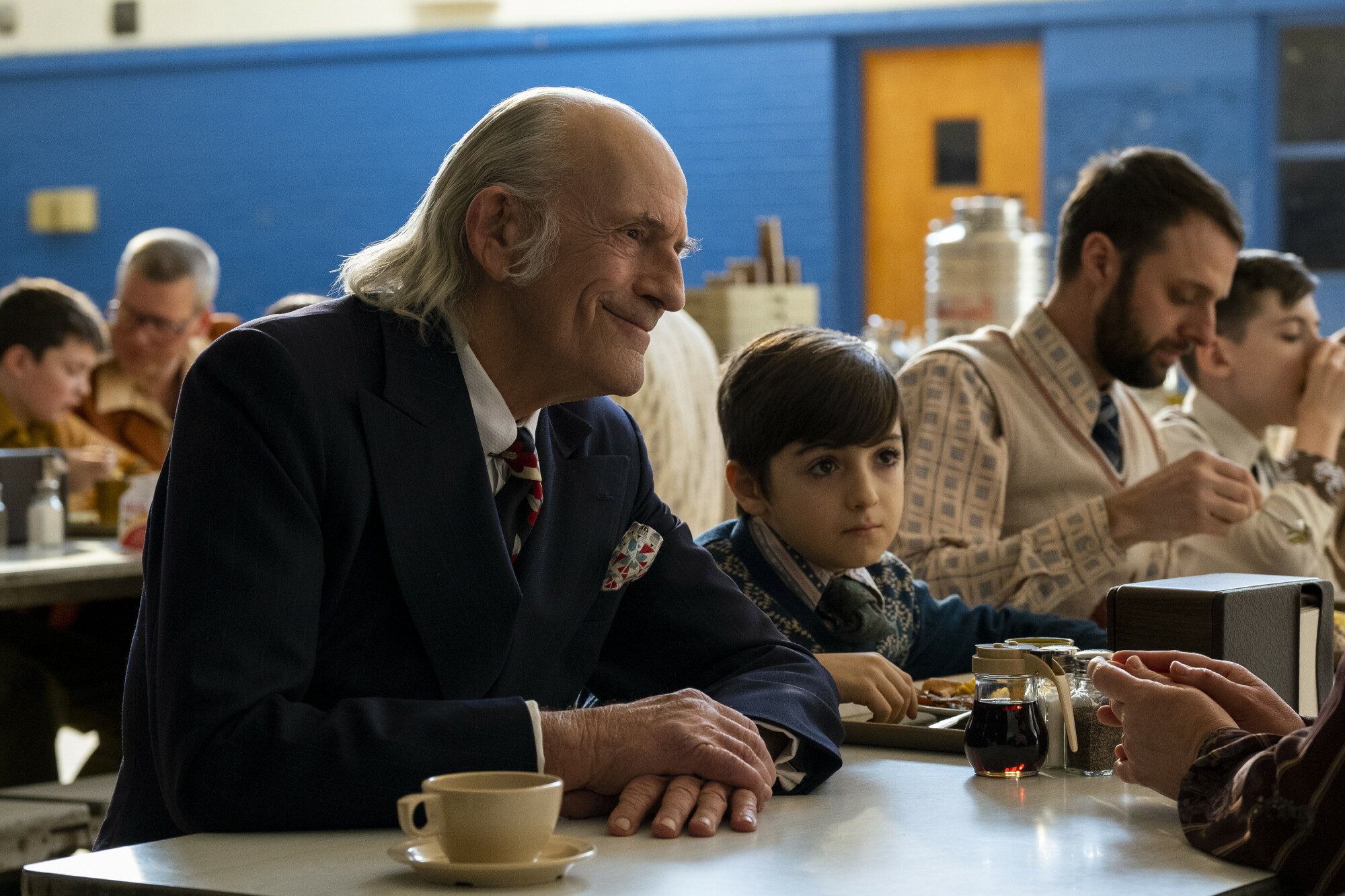 Christopher Lloyd in a suit sits next to a child at a cafeteria table in "The Tender Bar." 