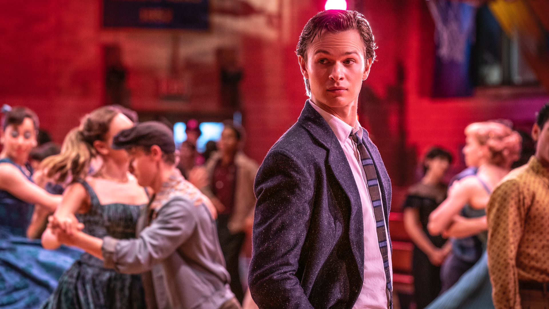 Ansel Elgort stands in a blazer, while others dance around him in 'West Side Story.'
