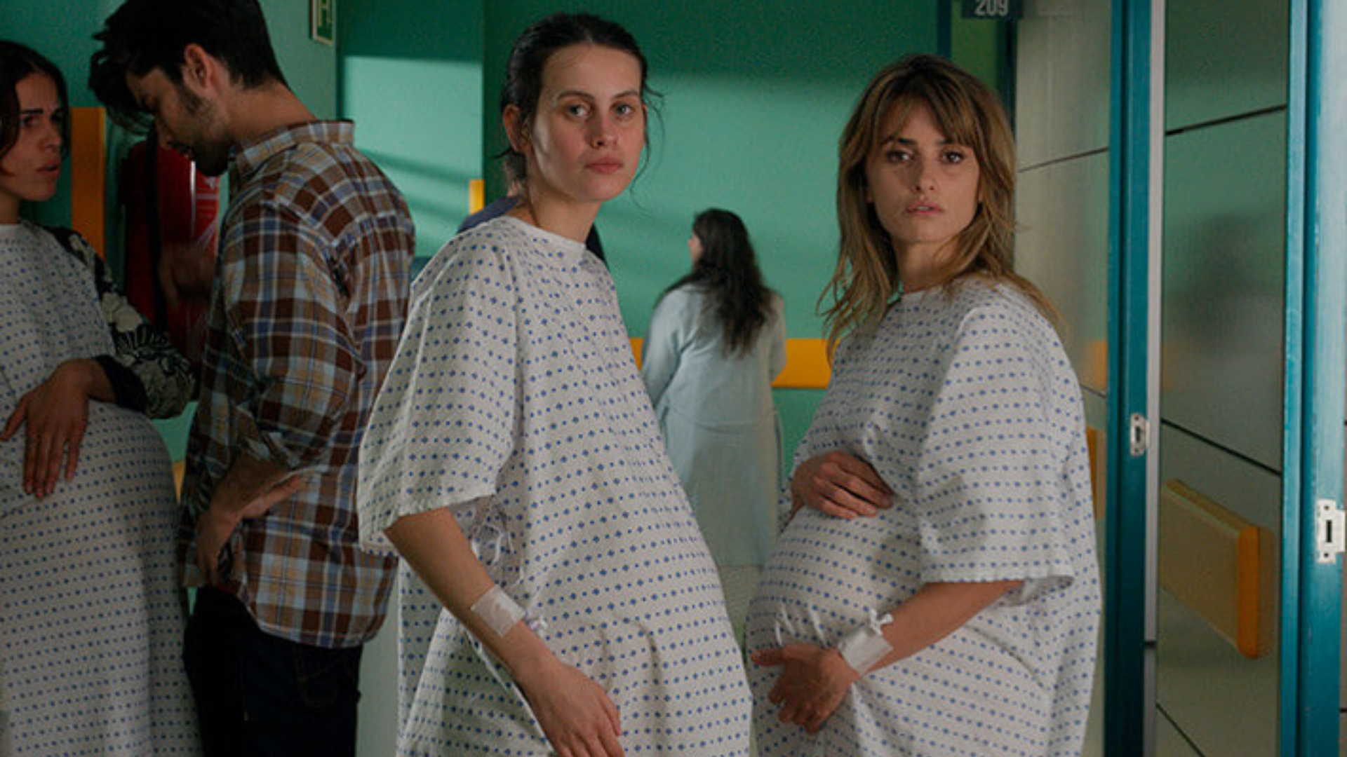 Two pregnant women in hospital gowns stand in a hospital hallway staring straight at the camera.