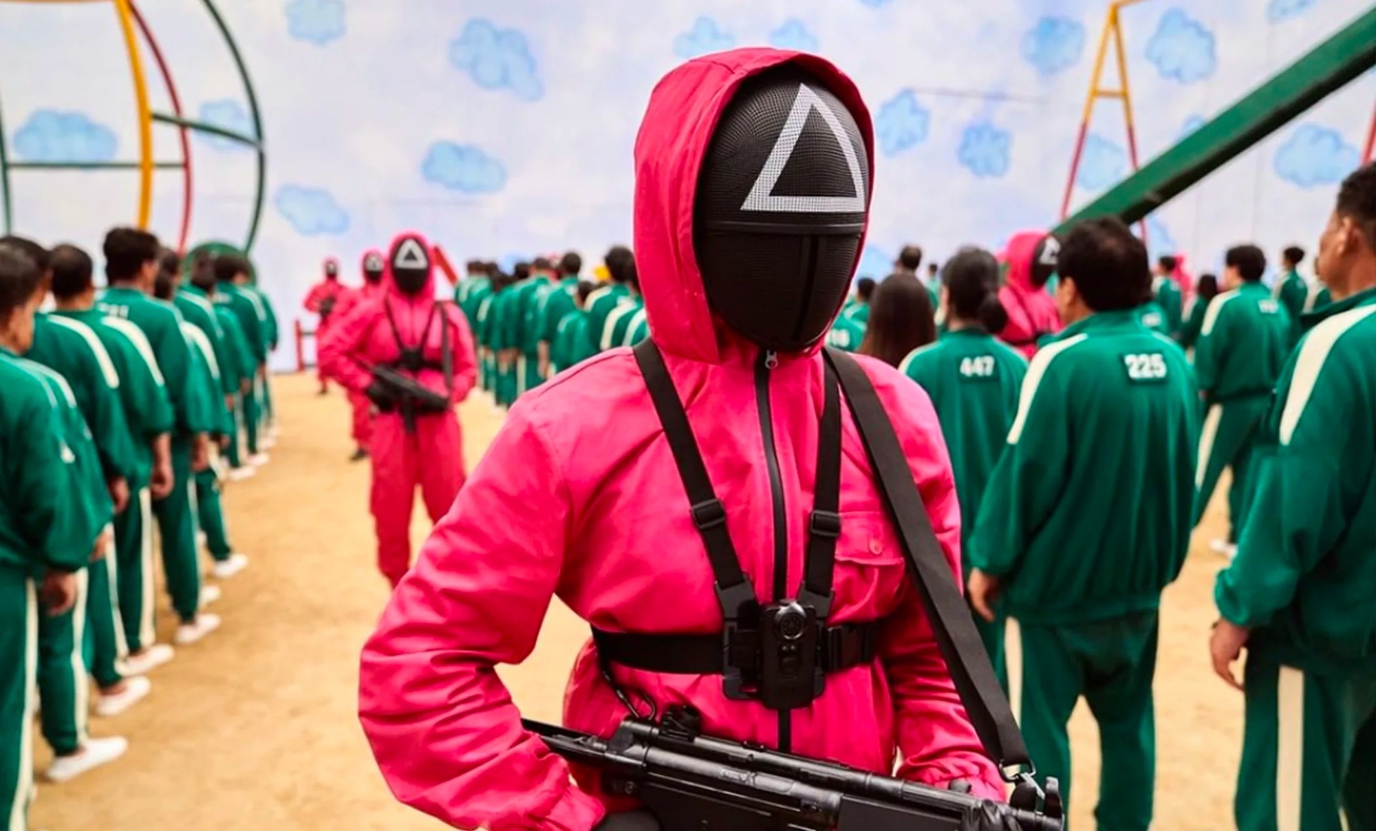 a person in a pink jumpsuit with a black mask with a white triangle on the mask carries a gun amongst contestants in green jumpsuits