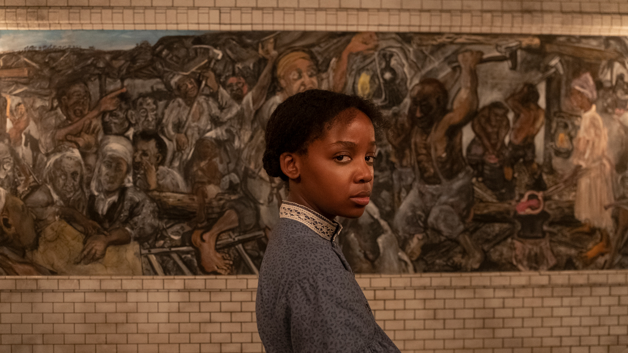 A young woman in a blue dress stands in front of a mural.