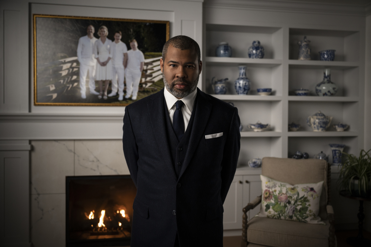 A man in a suit stands in a living room looking into the camera.