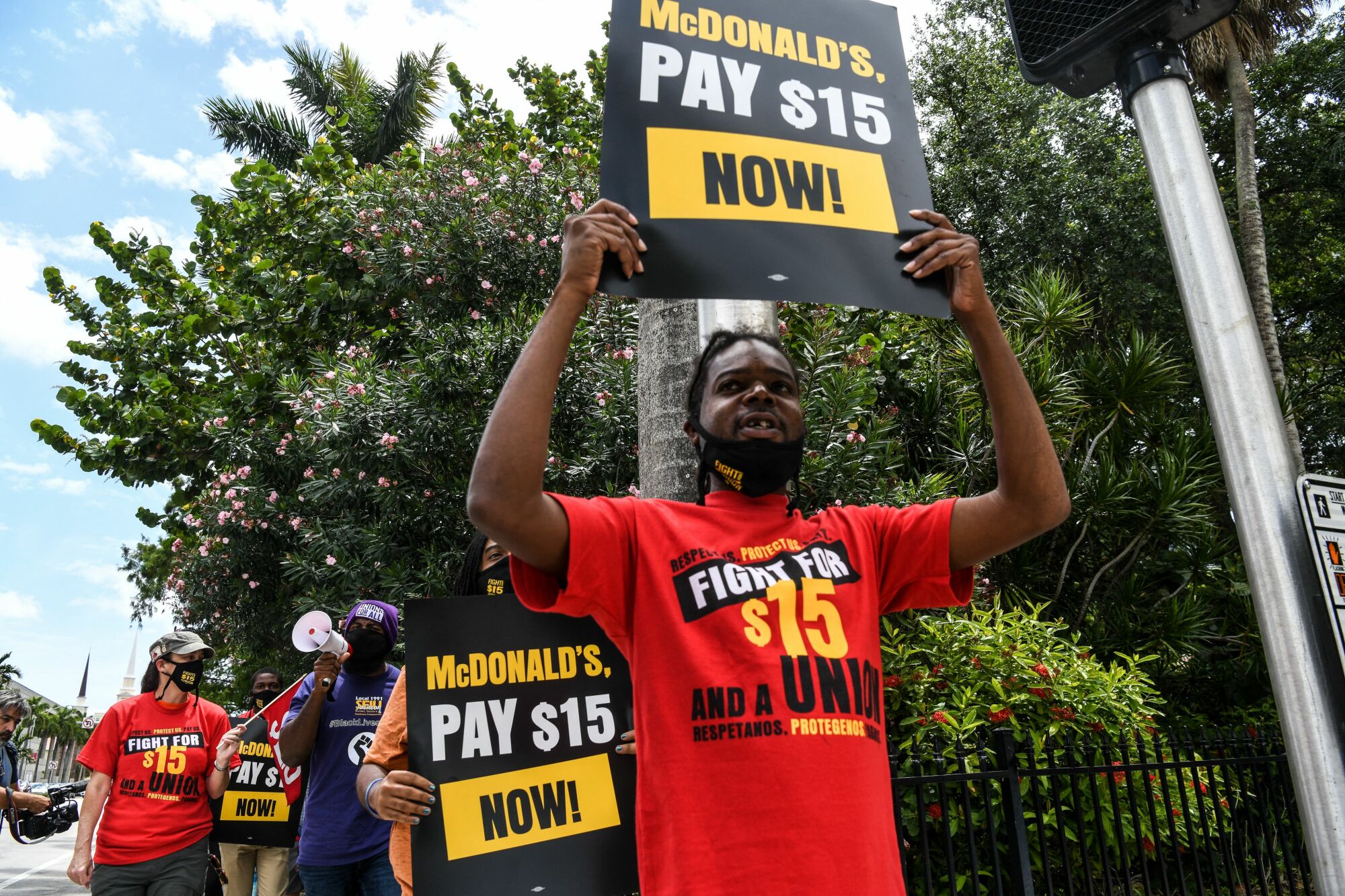 A man wears a red shirt that reads, "Fight for $15 and a union." He also carries a black sign that reads, "McDonald's, pay $15 now!"
