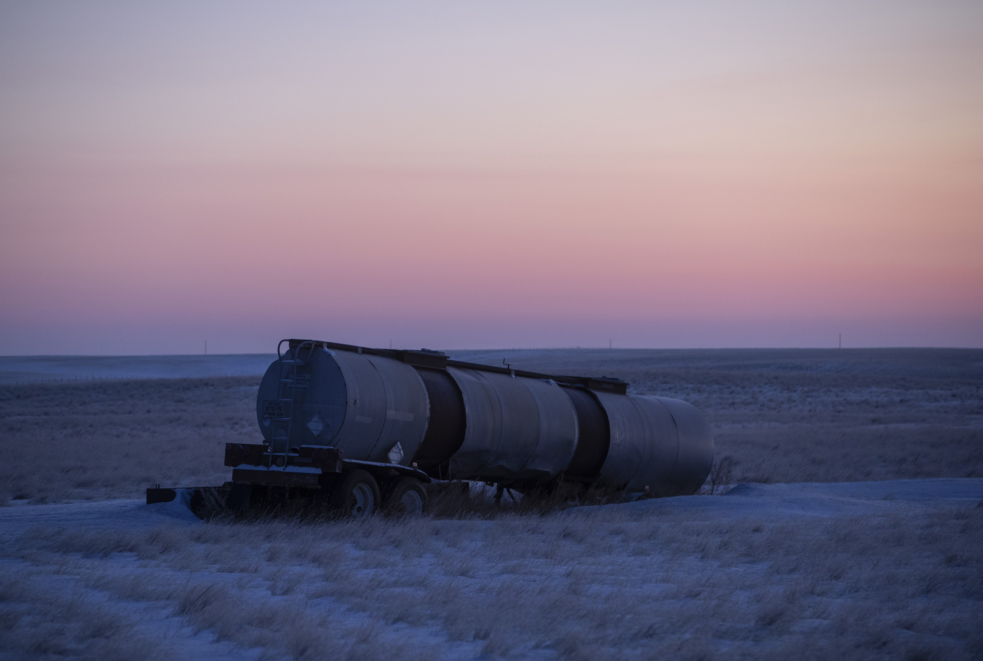 The sun rises behind an abandoned oil tanker truck parked awkwardly on a grass field.