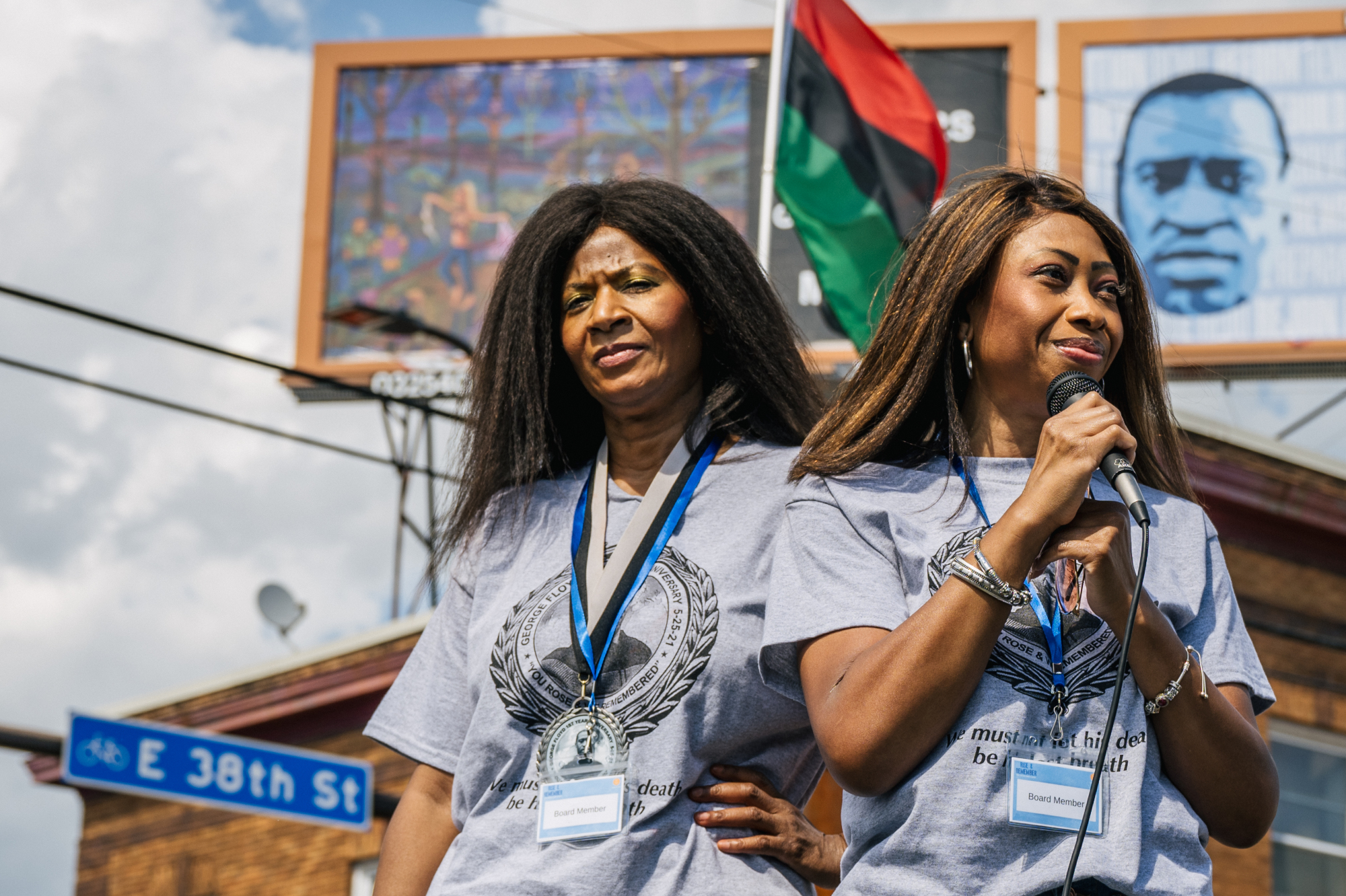Two women in gray shirts, one holding a microphone, speak to a crowd in front of a George Floyd memorial billboard.