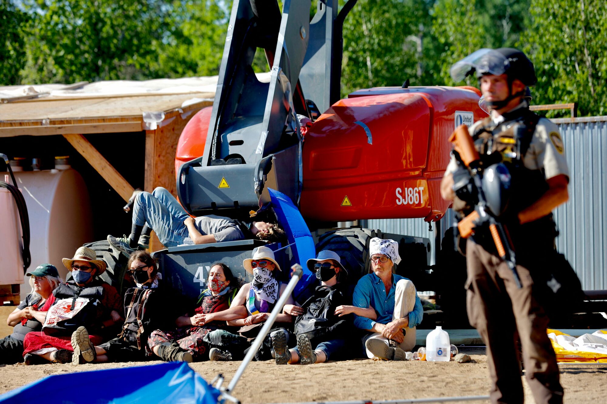 A police officer in riot gear holding a gun stands in the foreground as a group sits behind him in front of construction equipment.