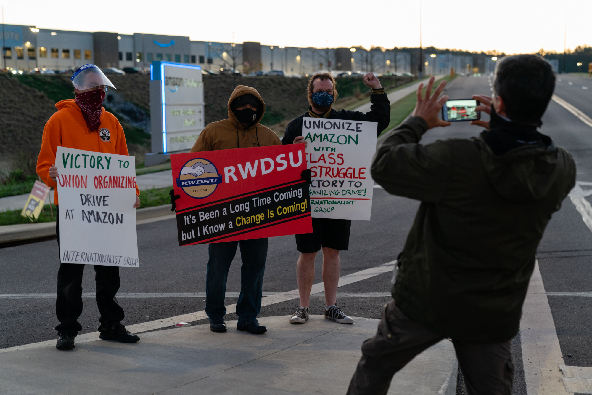 A person takes a picture of three Amazon workers holding signs asking for unionization.