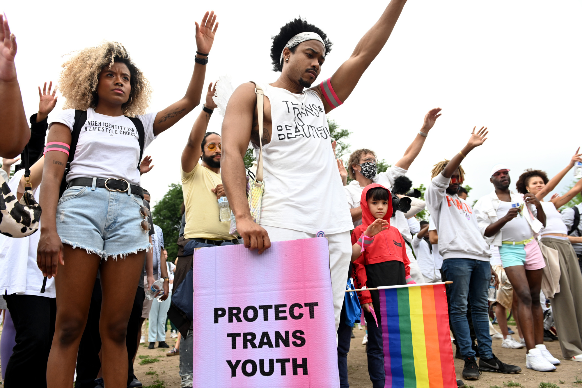 A photo of demonstrators standing in a line with their left hands raised. One holds a sign that reads, "Protect trans youth."