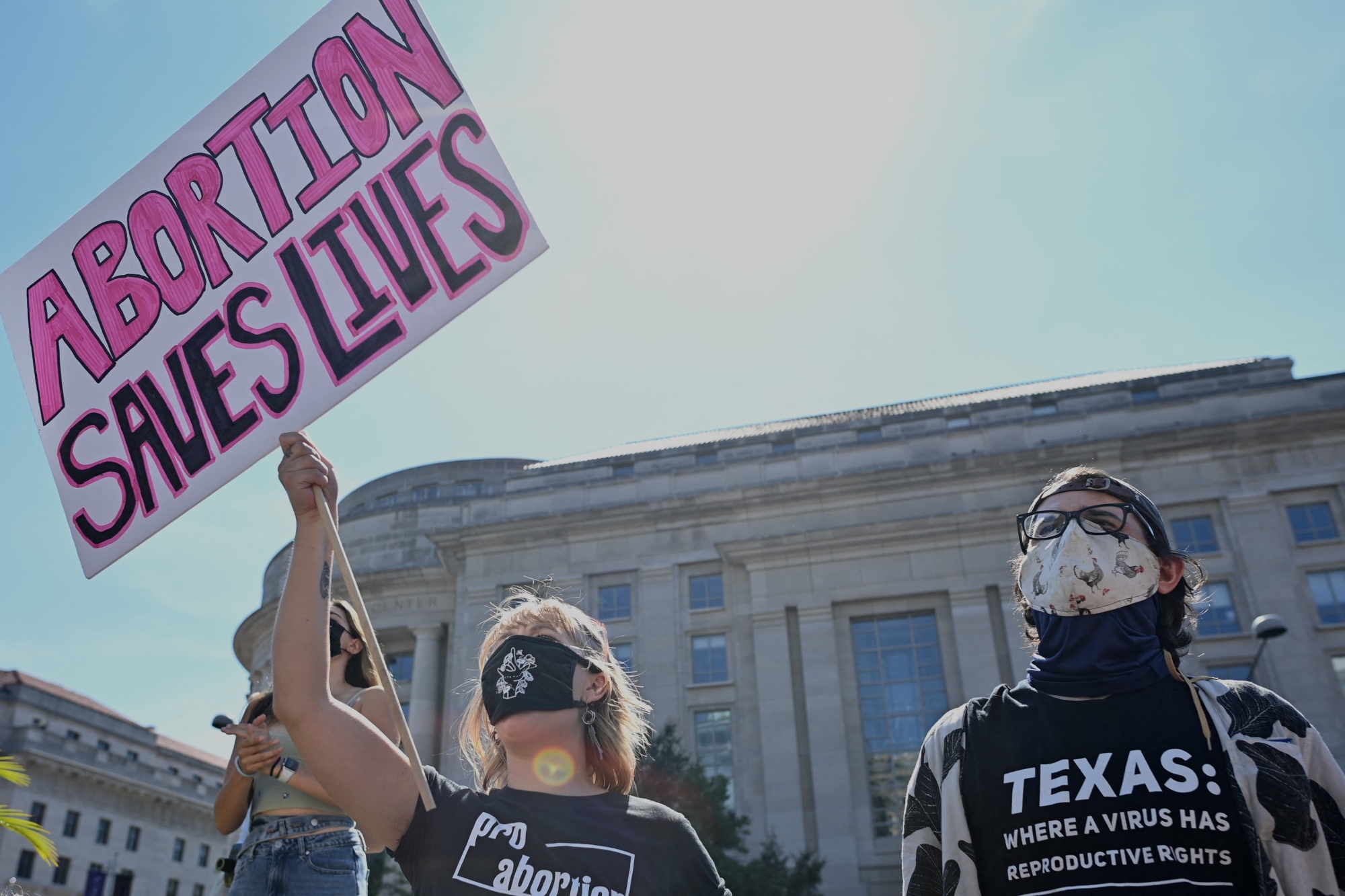A person in a mask stands next to another person holding a sign that reads, "Abortion saves lives."