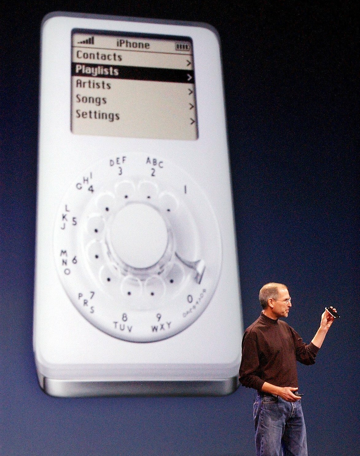 Steve Jobs stands in front of a spoof image of an iPod with a rotary dial, pretending it's the iPhone. 