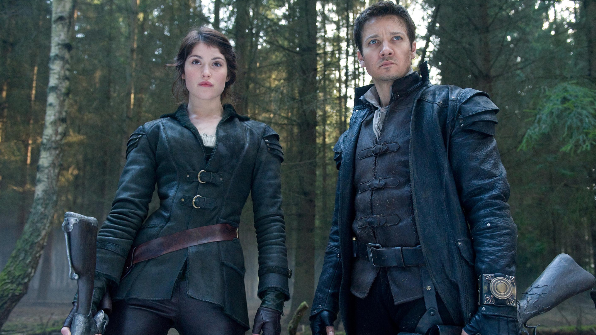 A man and woman in black leather armor stand in front of a forest. Both carry large firearms. 