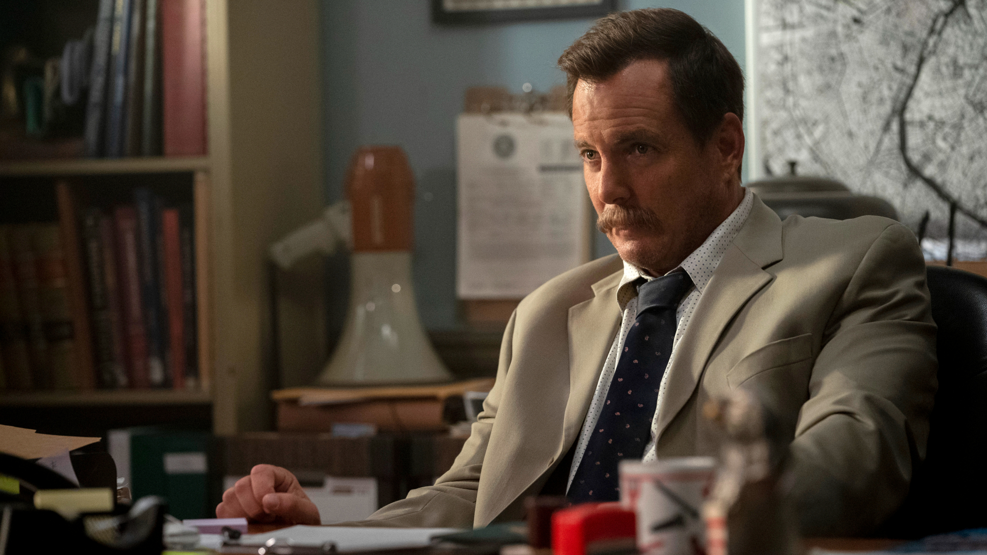 A mustachioed man sits at a desk