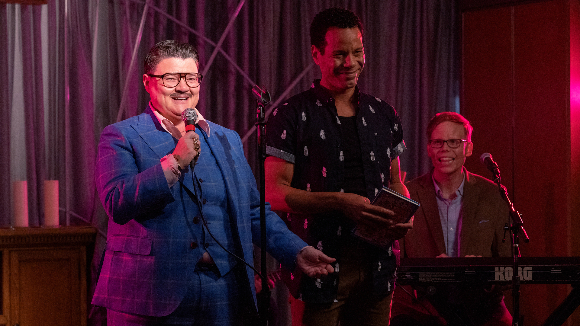 Three men on stage. One holds a microphone, one sits behind a keyboard.
