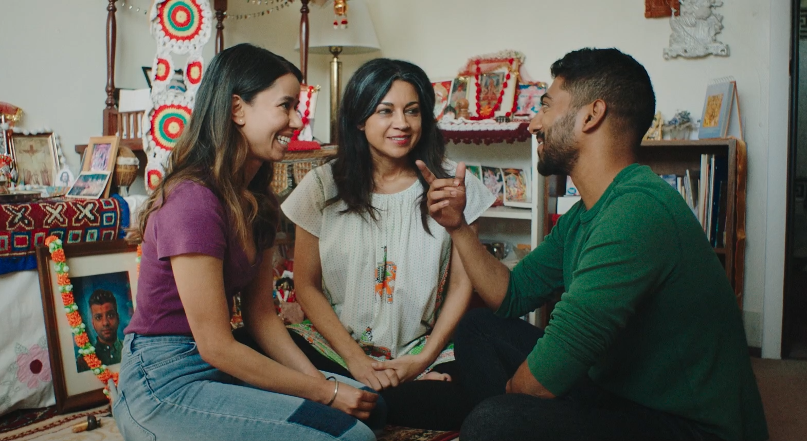 A woman sits on the floor with her adult children as they all smile and talk.