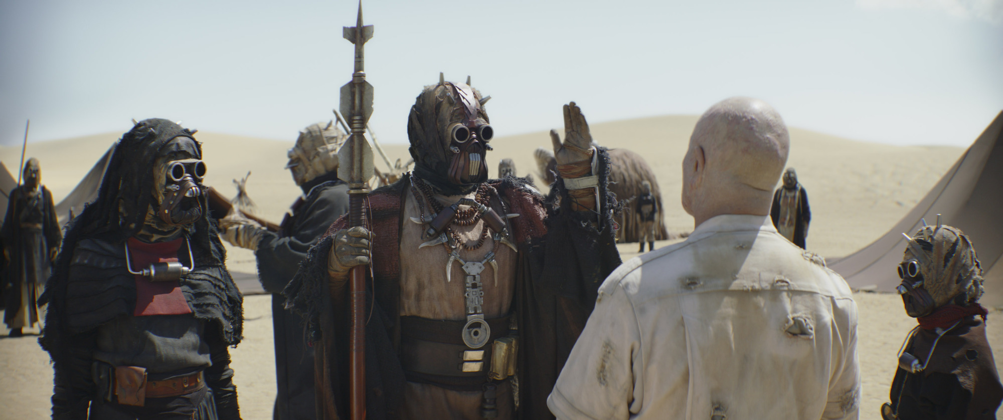 A still from "The Book of Boba Fett" featuring Boba Fett, sans armor and facing away from the camera, as he talks to a group of robed and masked Tusken Raiders.