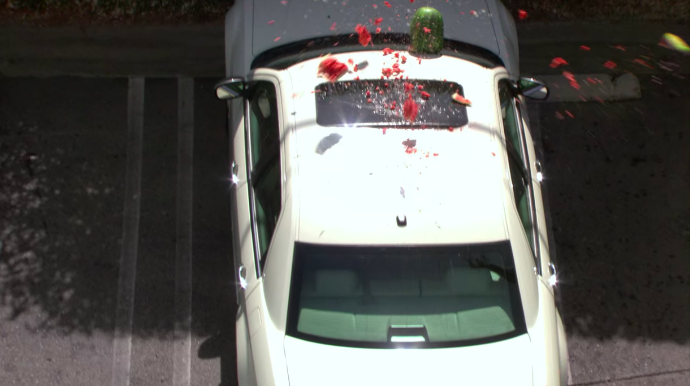 A watermelon smashing on the top of a white Chrysler 300 in a parking lot.