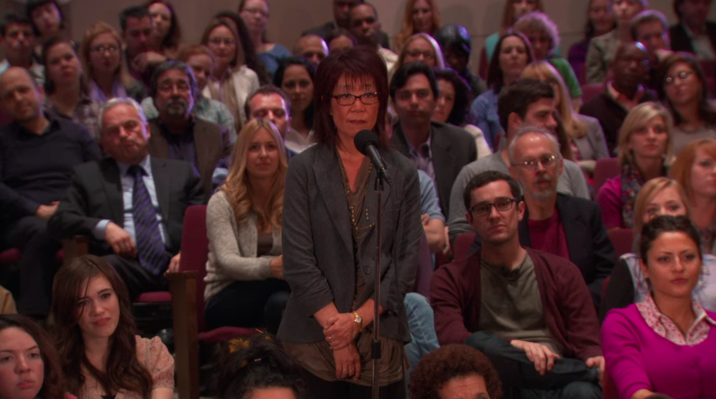 A crowd of extras sitting in auditorium seats during "The Office" finale. A woman, OfficeTally creator Jennie Tan, is standing in front of a microphone asking a question.