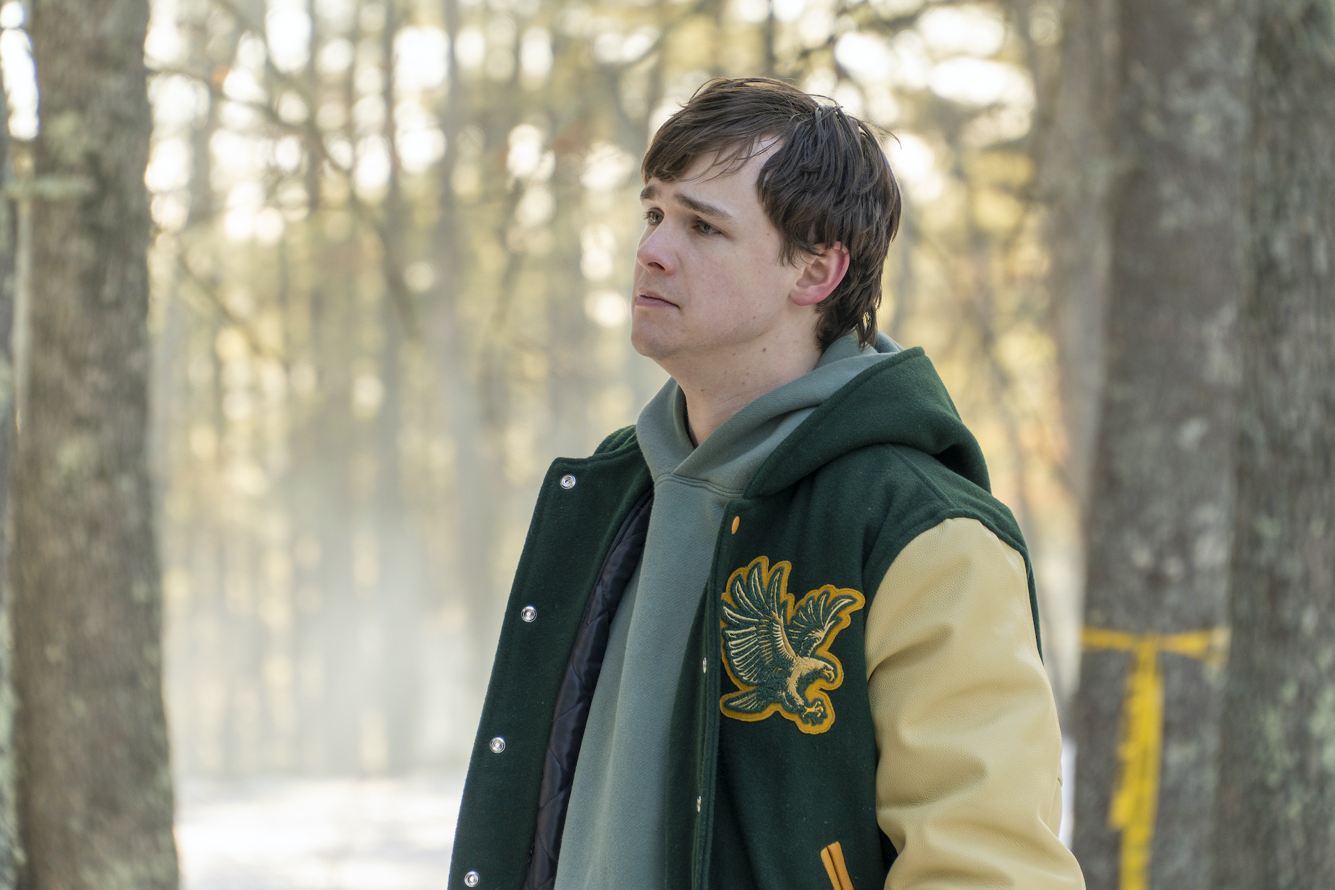 A high school boy in a letter jacket looks sad while standing in a snowy forest.