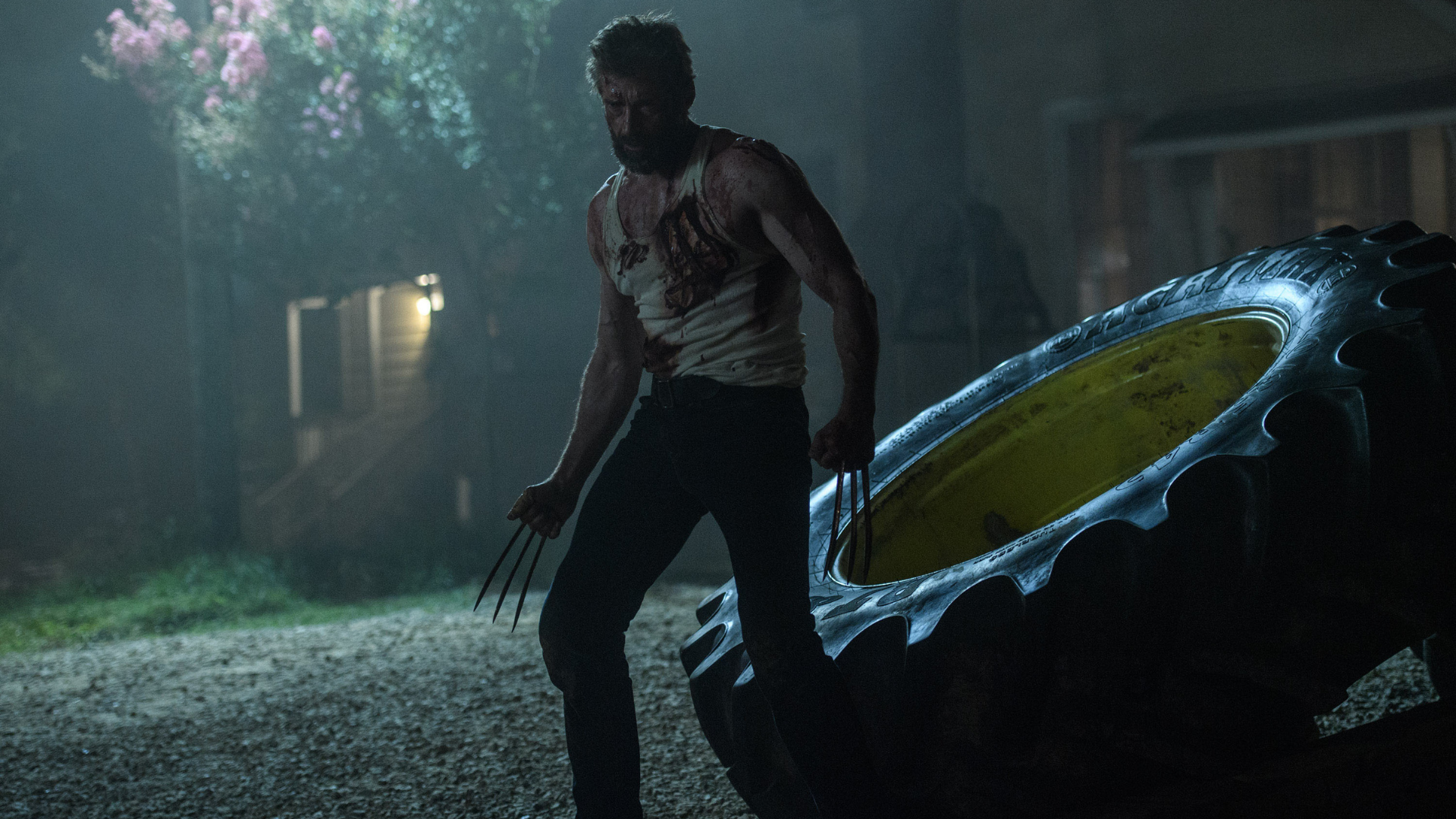 A man with claws protruding from his fists stands in front of a farm house at night. 