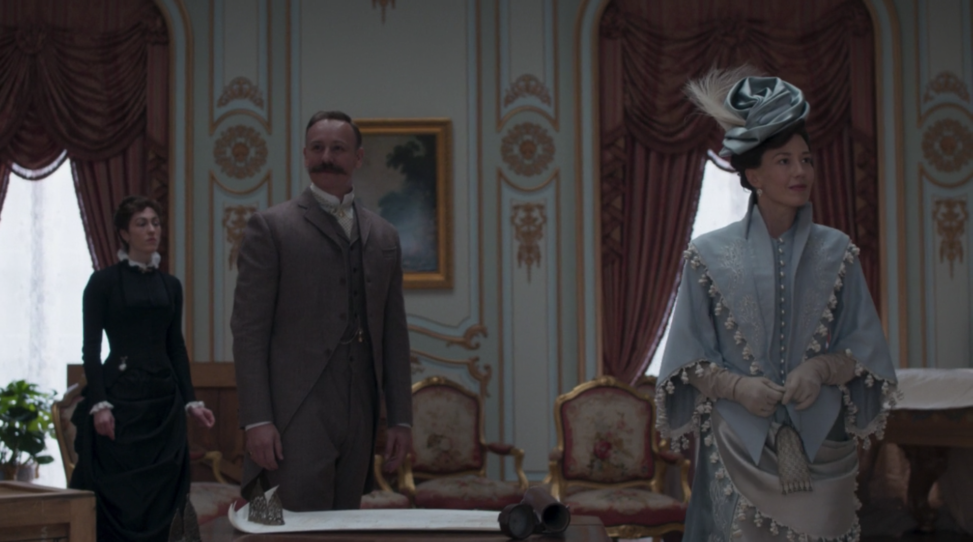 A woman in a light blue 19th century dress and matching cape walking through an opulent room.