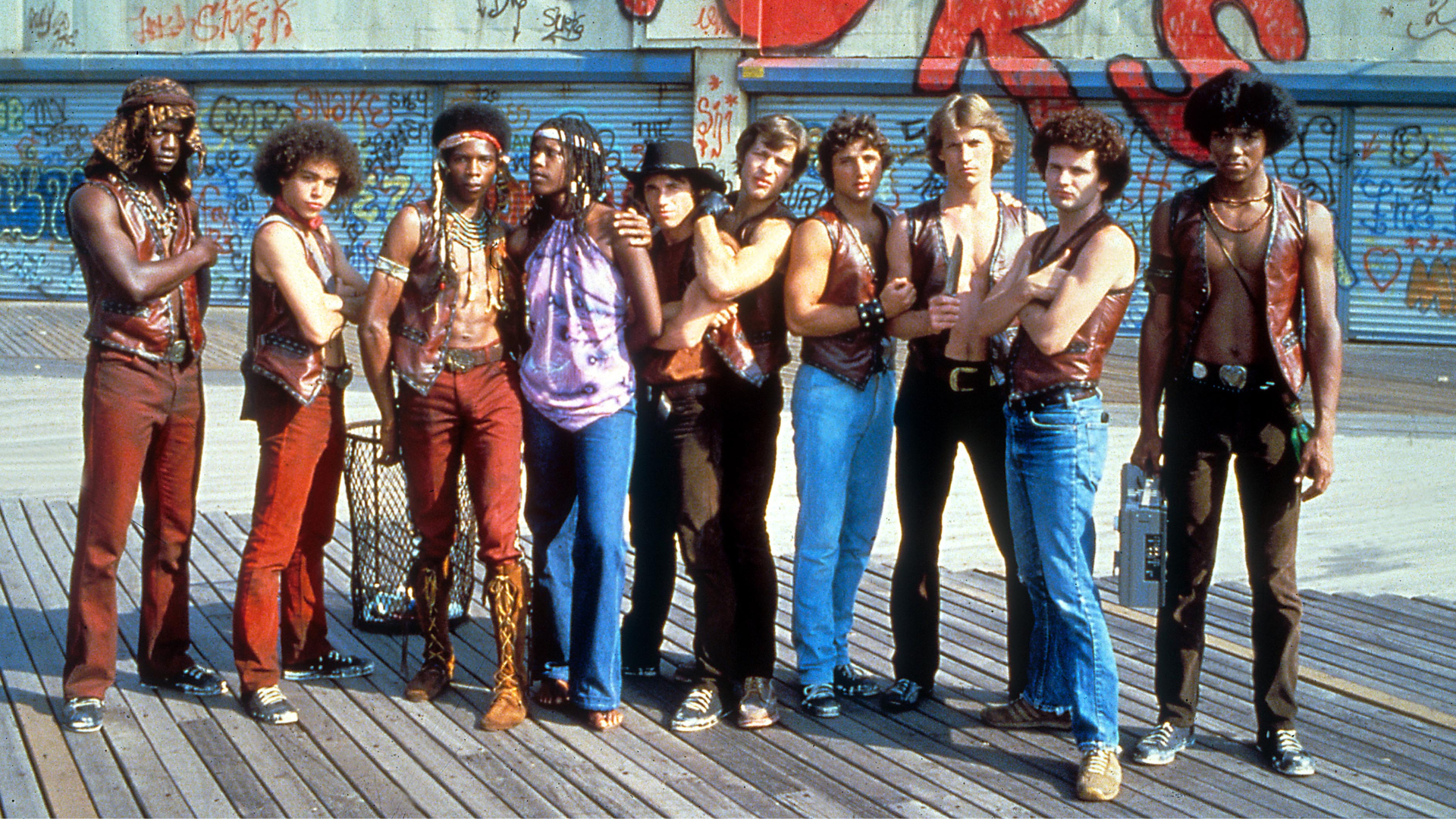 A gang of young men stand on the Coney Island boardwalk. All are dressed in red slacks and leather vests with no shirts. 