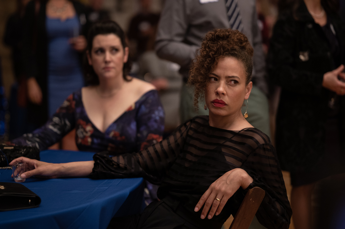 Two middle-aged women in cocktail dresses, sitting at a table for their high school reunion.