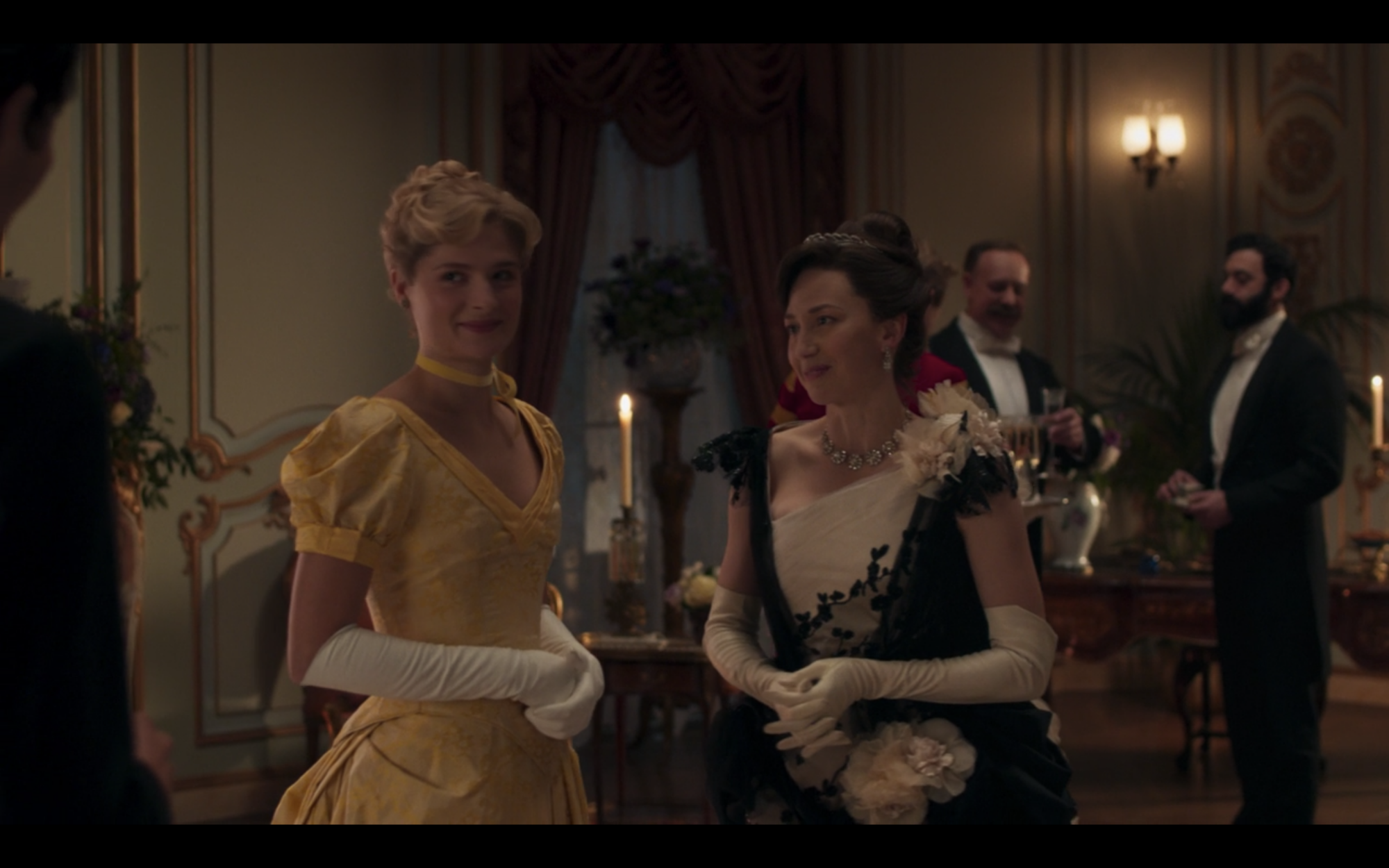 Two women, a blonde on the left in a yellow dress and a dark-haired one on the right in a black-and-white 19th century ball gown