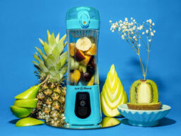 Blue portable blender with various fruit on the inside and pineapple, green apple, pear, and decorative kiwi beside it