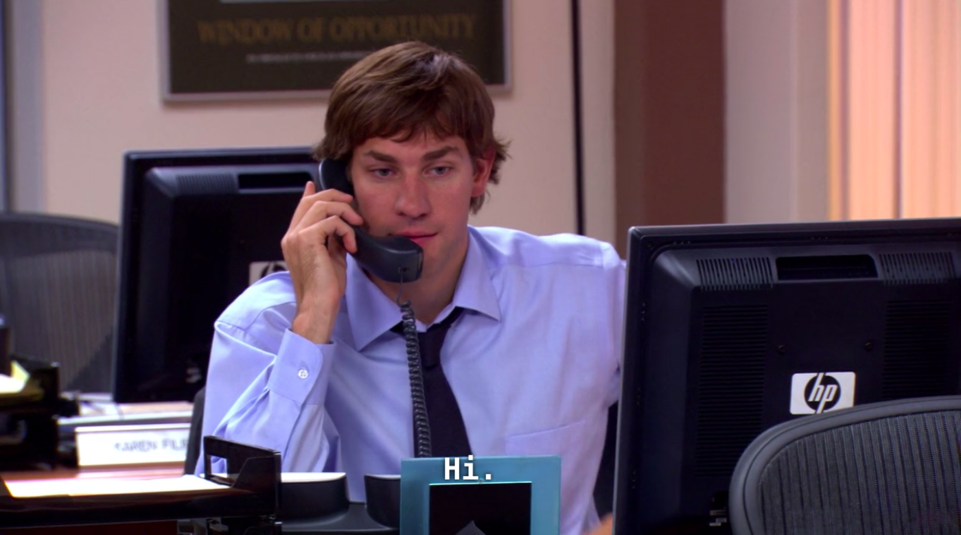 A man, John Krasinski (who plays Jim Halpert on "The Office"), sitting at a desk and holding a phone to his ear.