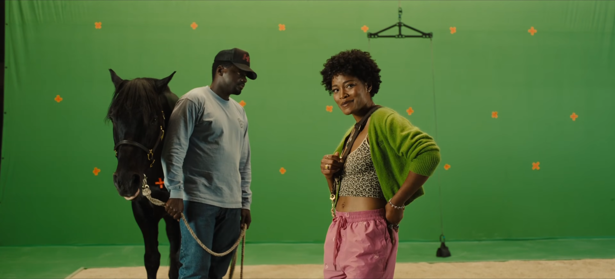 Two people, a man and a woman, stand in front of a green screen on a production strange. The man is standing in the background with a horse looking downward. The woman is staring directly at the camera and grinning.