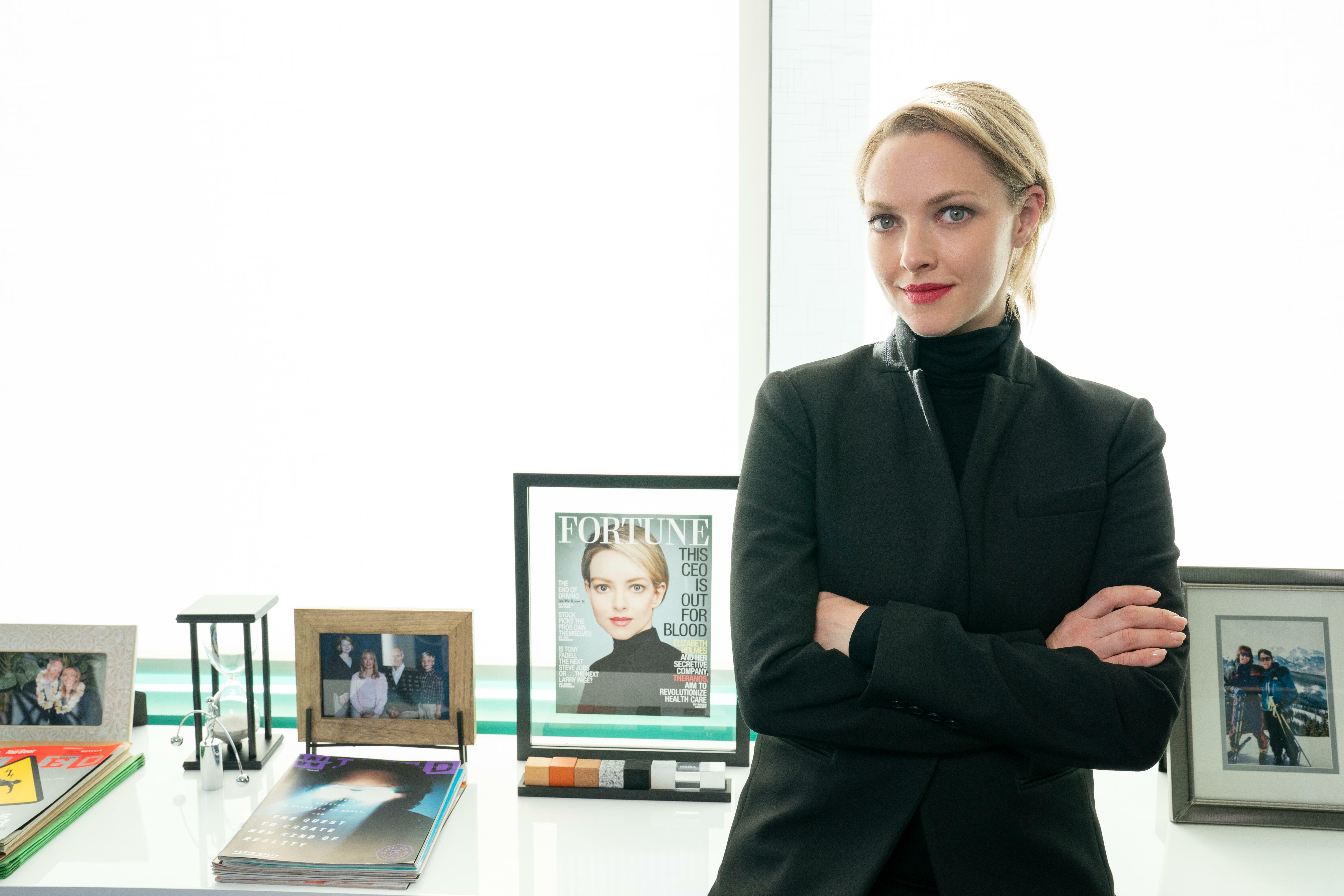 A blonde woman in a black blazer and turtleneck stands with her arms crossed