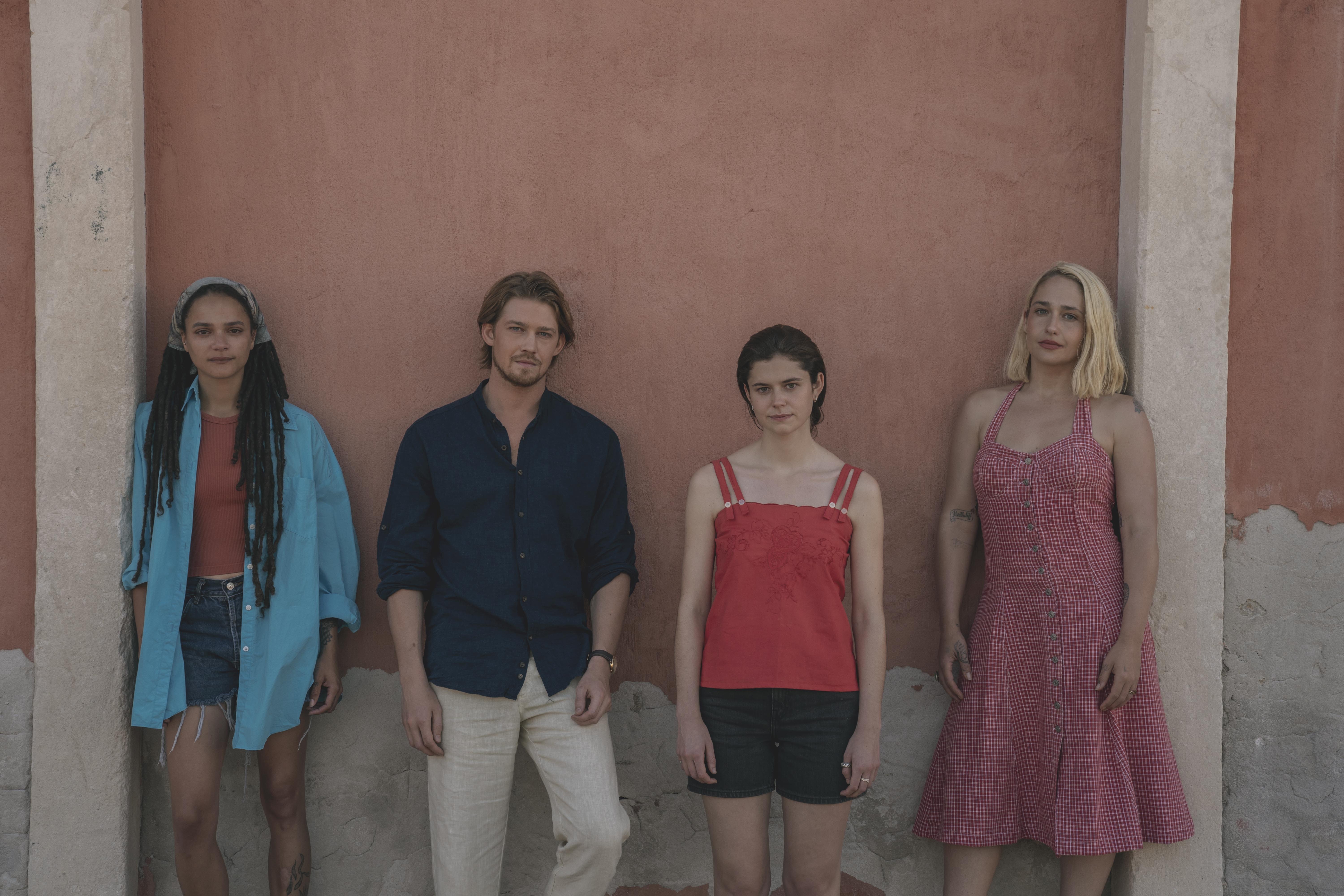 The cast of Conversations With Friends pose in front of an orange wall. Jemima Kirke plays Melissa, Joe Alwyn plays Nick, Alison Oliver plays Frances alongside Sasha Lane as Bobbi.