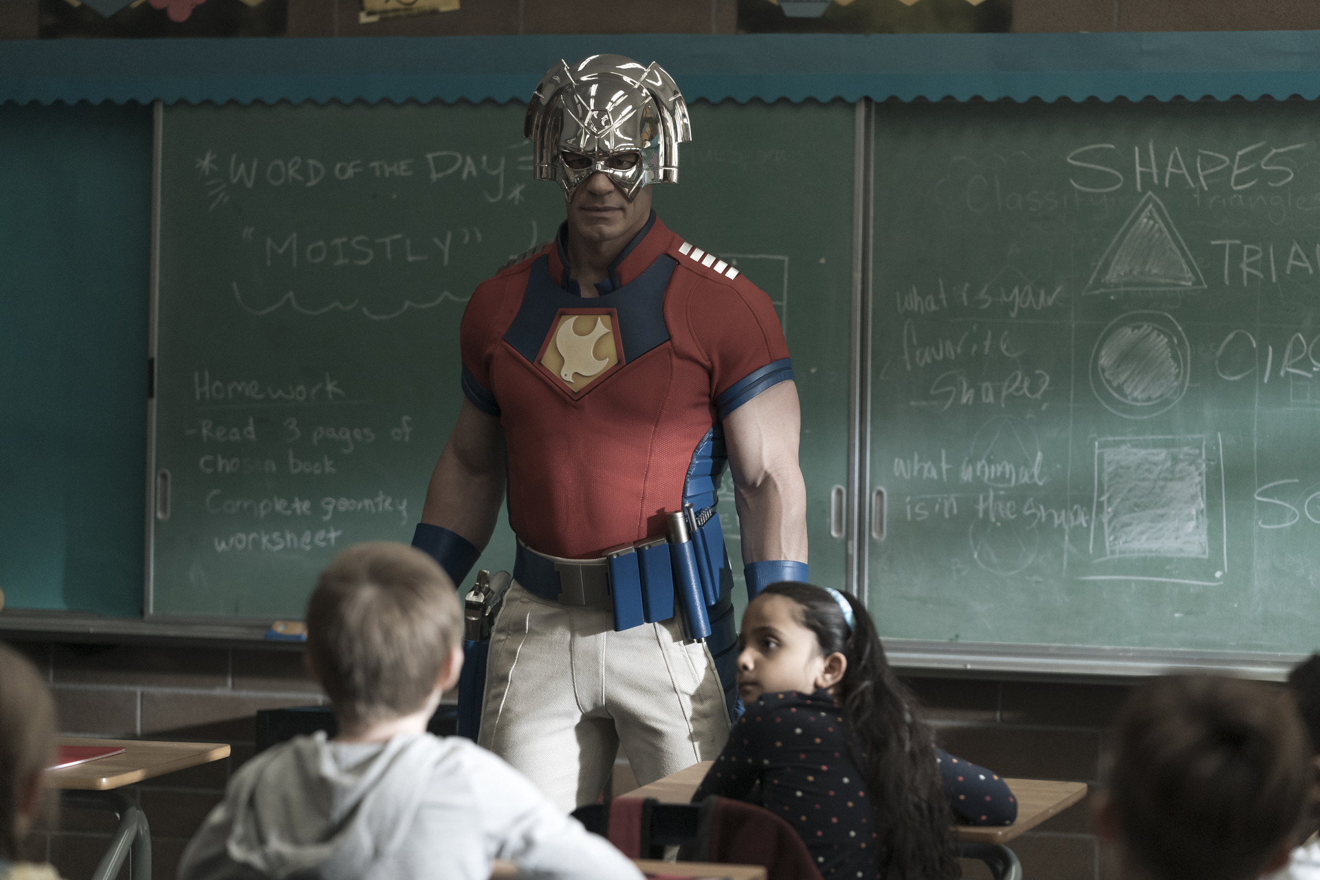 A man in a tight red superhero outfit and metal helmet addresses a classroom of children; a still from