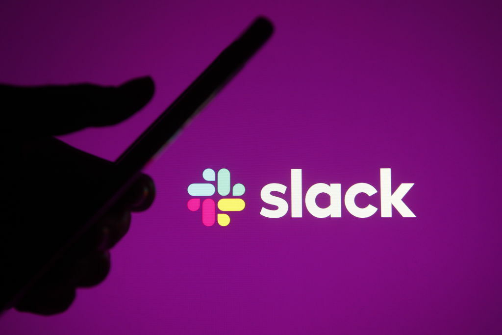 A photo of the Slack logo behind a silhouette of a hand holding a smartphone.