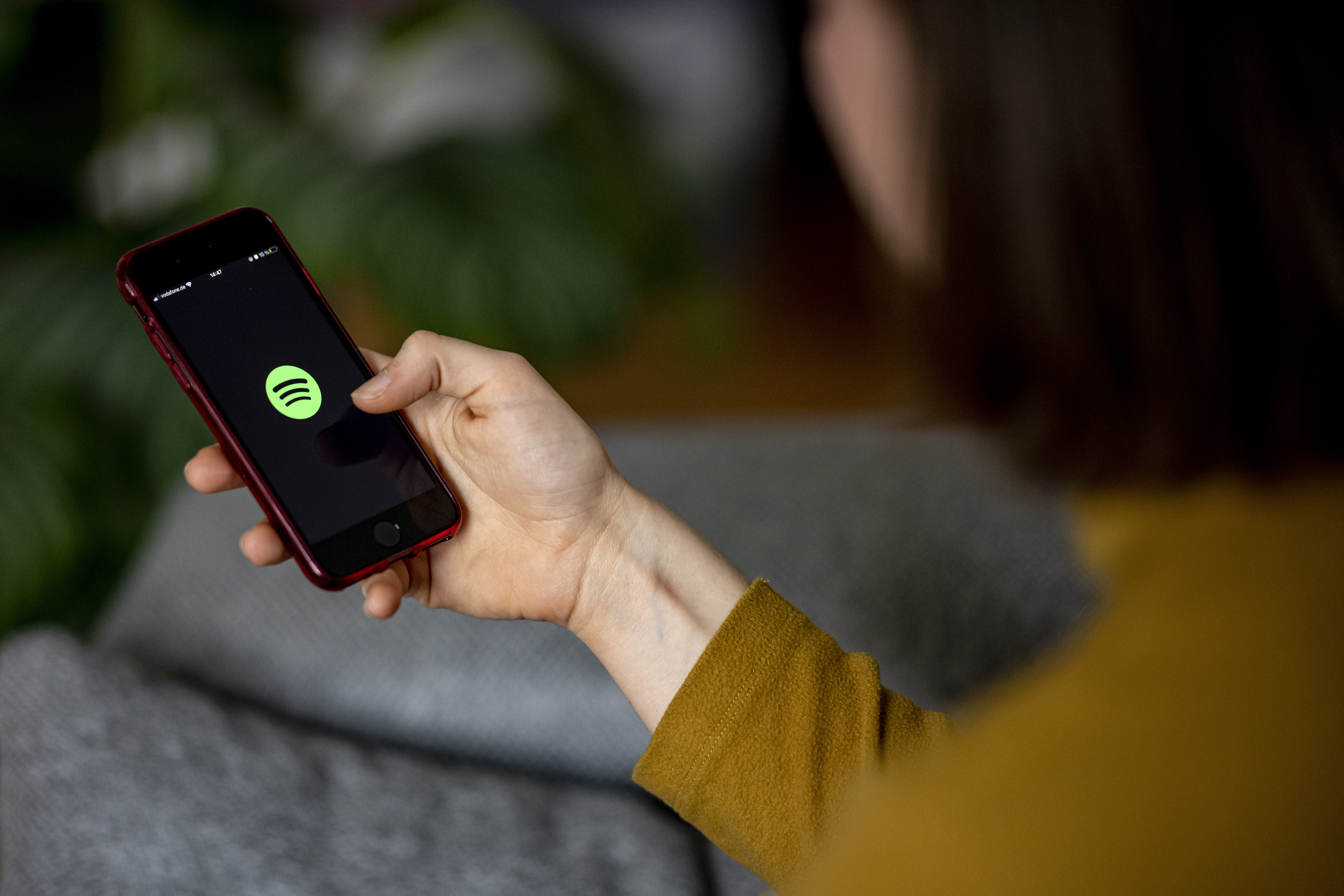 A woman's hand holds out a phone with the Spotify logo on the screen. She is wearing a yellow sweater. 