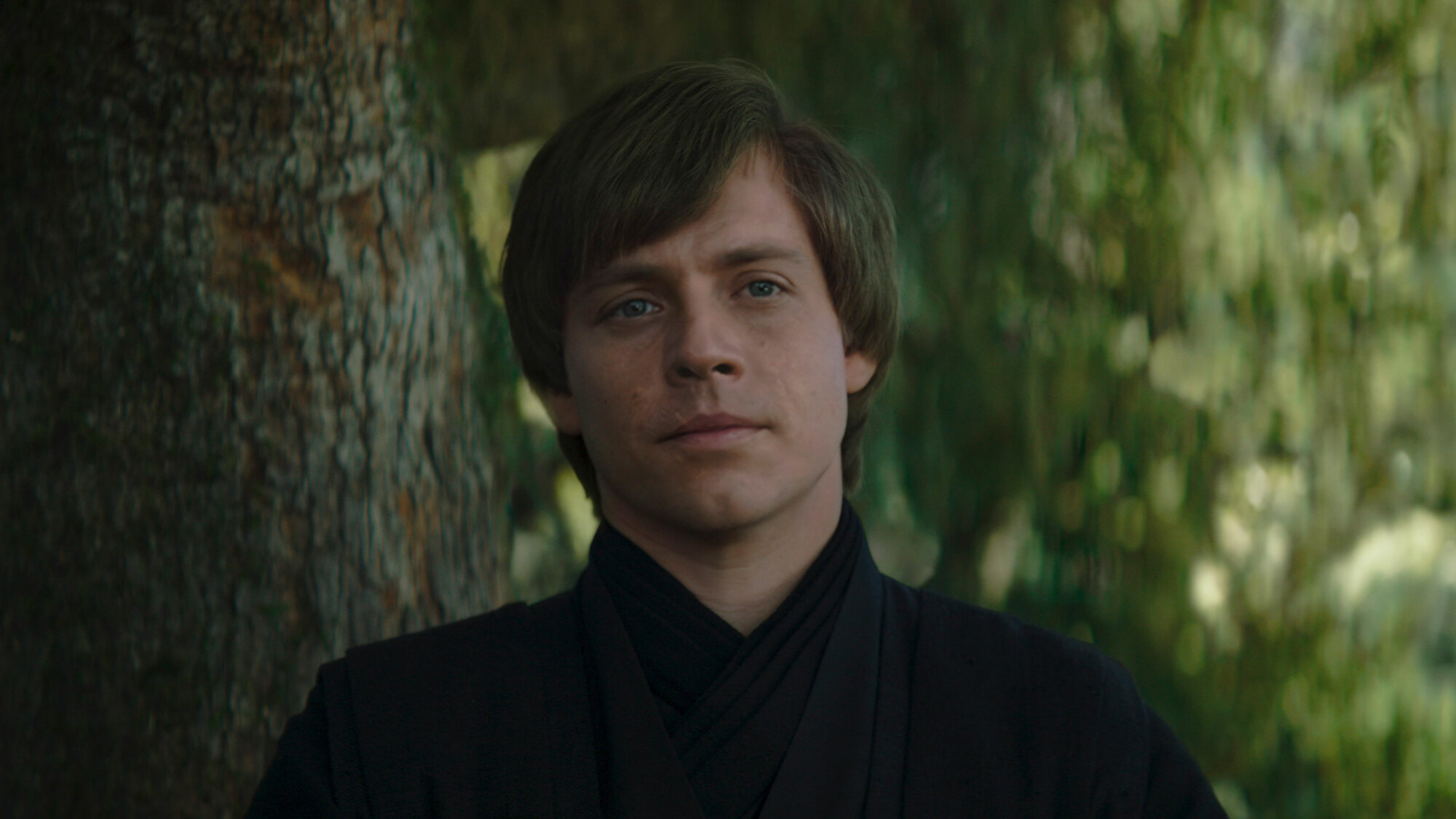 A close-up of a man in a forest setting. It's a still from "The Book of Boba Fett" and the man is a digital recreation of Mark Hamill's younger Luke Skywalker.