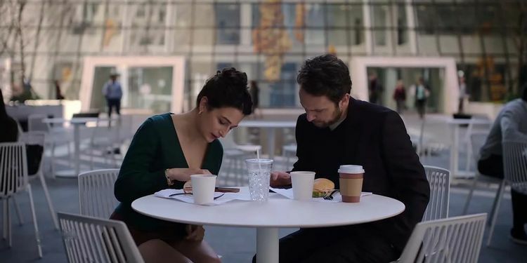A woman (Jenny Slate as Emma in "I Want You Back") and a man (Charlie Day as Peter) sitting at a table together while eating food and using their phones.