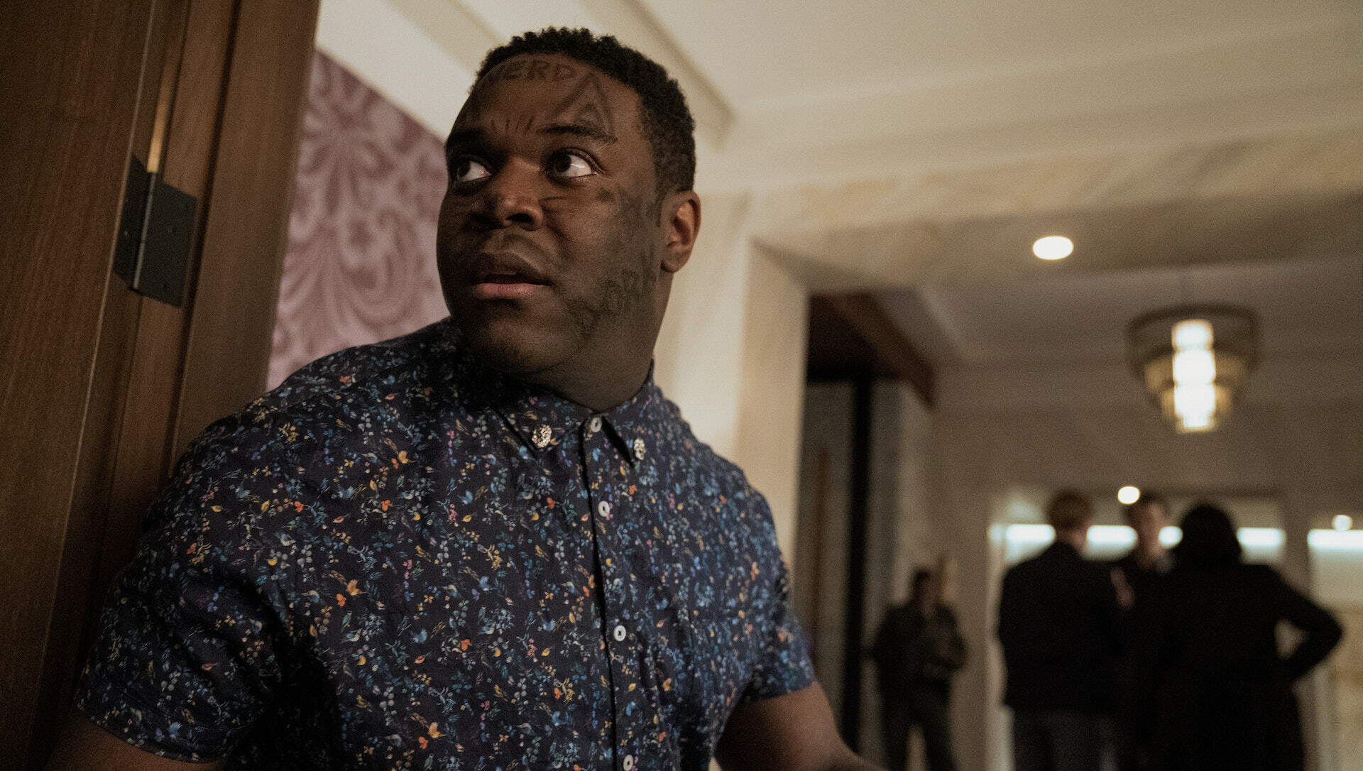 A man with drawings on his face looks around nervously while police line the background; a still from "The Afterparty."
